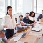 A businesswoman standing at a conference table explaining something to many business people