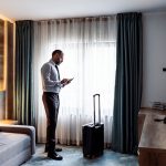A man standing by the window in a hotel room with a suitcase next to him
